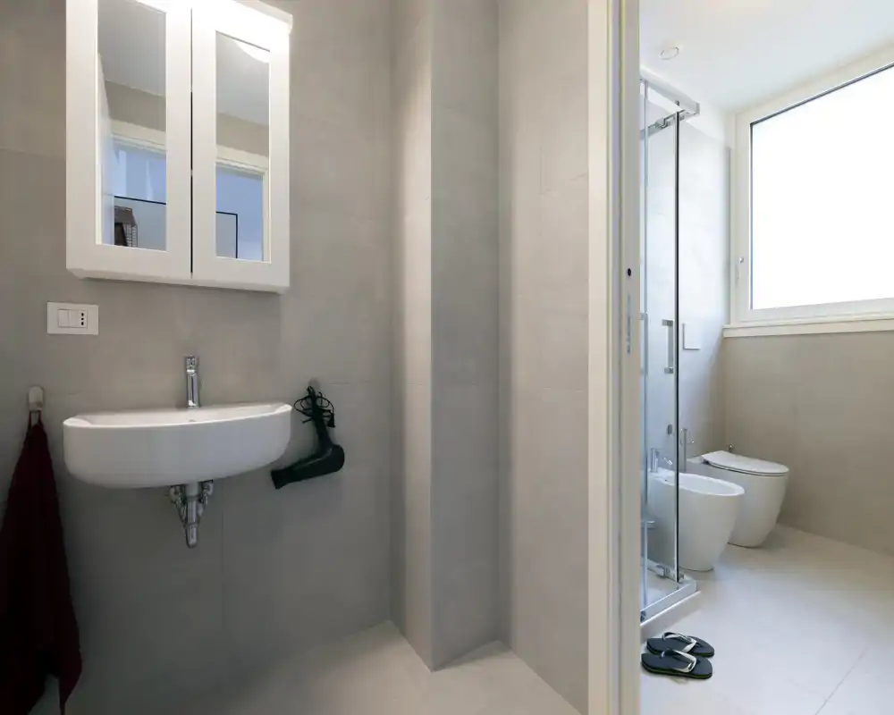 Modern, bright bathroom with a wall-mounted sink, mirror cabinet, shower enclosure, bidet and toilet; neutral gray tiles and a large frosted window provide plenty of natural light.