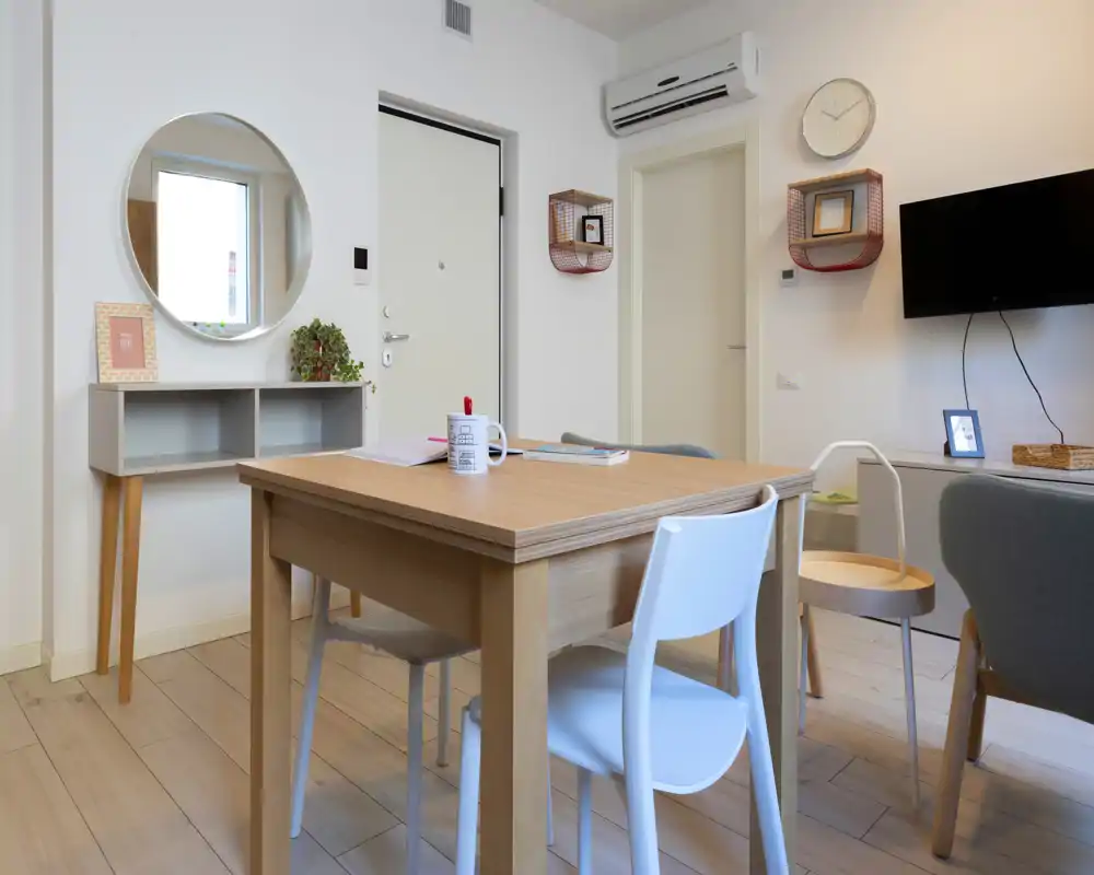 Bright, modern living/dining area with a wooden table, mixed chairs, wall-mounted TV and tasteful decor — ideal for a compact apartment.