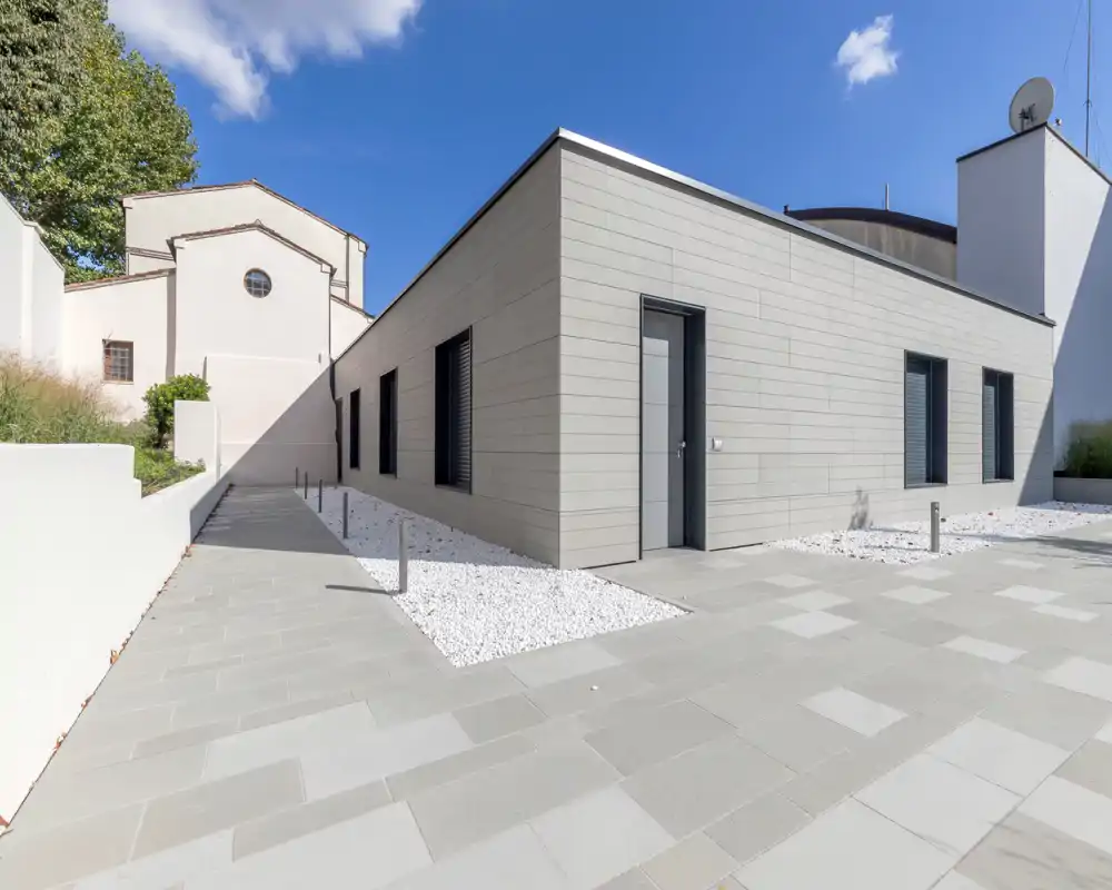 Modern exterior courtyard with paved walkway and minimalist landscaping featuring white gravel and a single-story building facade under a bright blue sky.