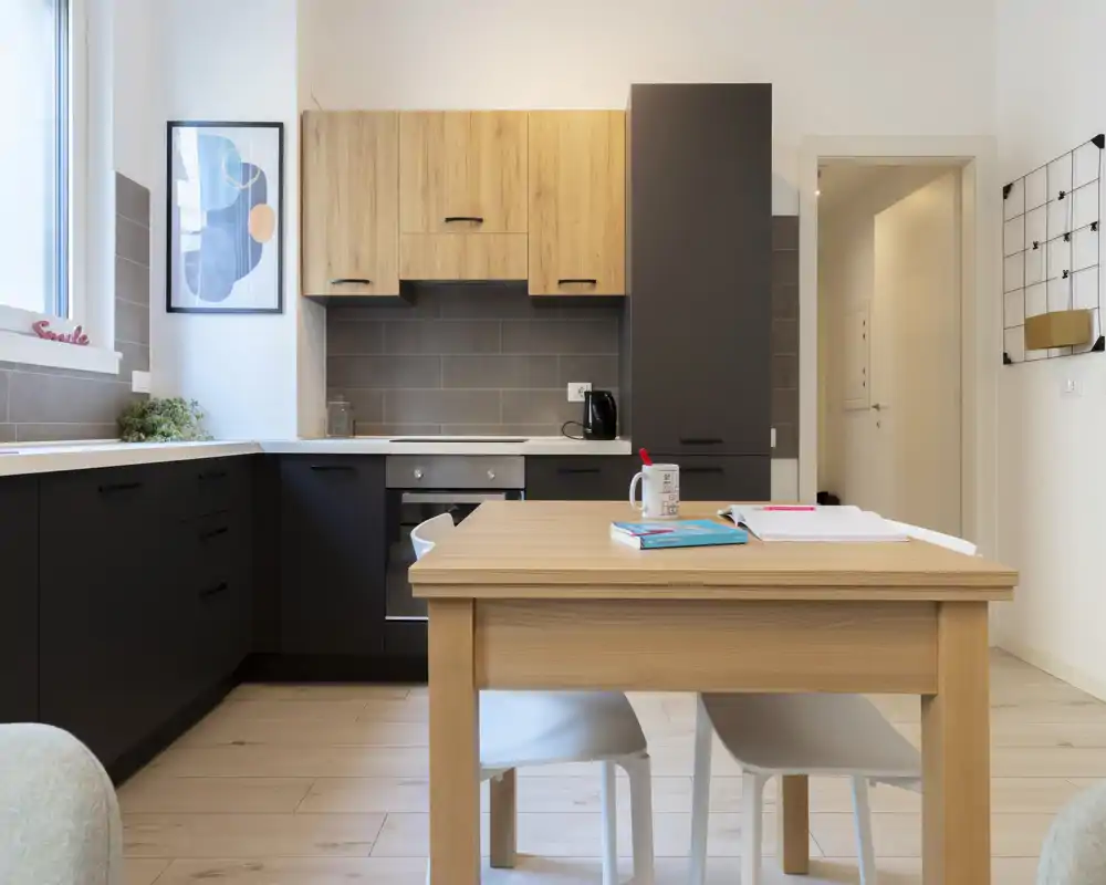 Modern compact kitchen with a wooden table in the foreground, matte dark cabinets, light wood upper units and a built-in oven. Bright, tidy space with simple decor ideal for short-term rentals.