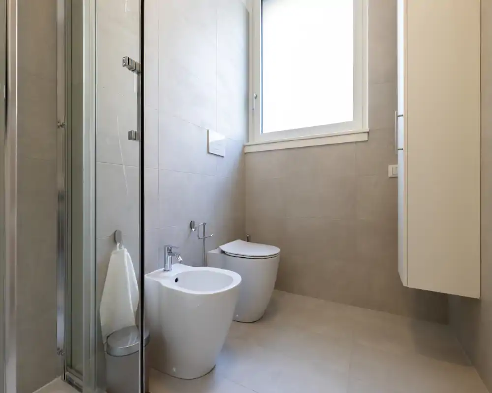 Bright, modern bathroom with a wall-mounted toilet, bidet and a glass shower screen; neutral tiles and a high window create an airy, minimal look.