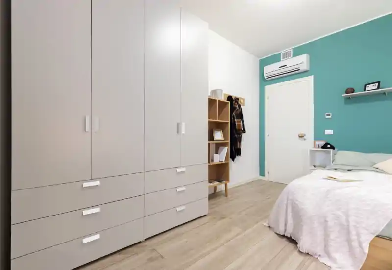 Bright, modern bedroom with a large wardrobe, pale wood floor and a neatly made bed with soft pink throw. The room features clean lines, a teal accent wall, shelving and an air-conditioning unit for comfort.