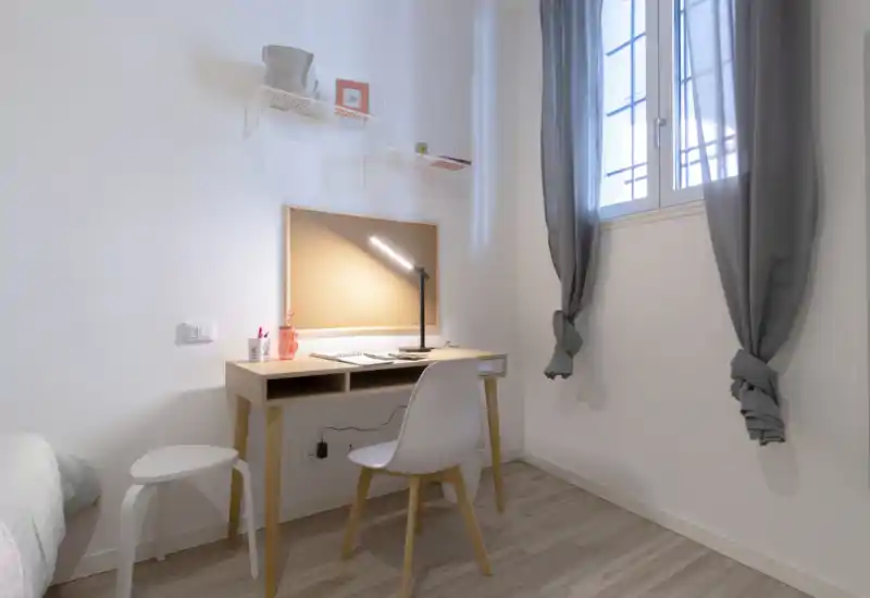 Small, bright bedroom corner with a minimalist wooden desk, chair and a bedside visible at the left edge. Natural light from a tall window, neutral decor and a focused desk lamp create a clean study nook within the sleeping area.