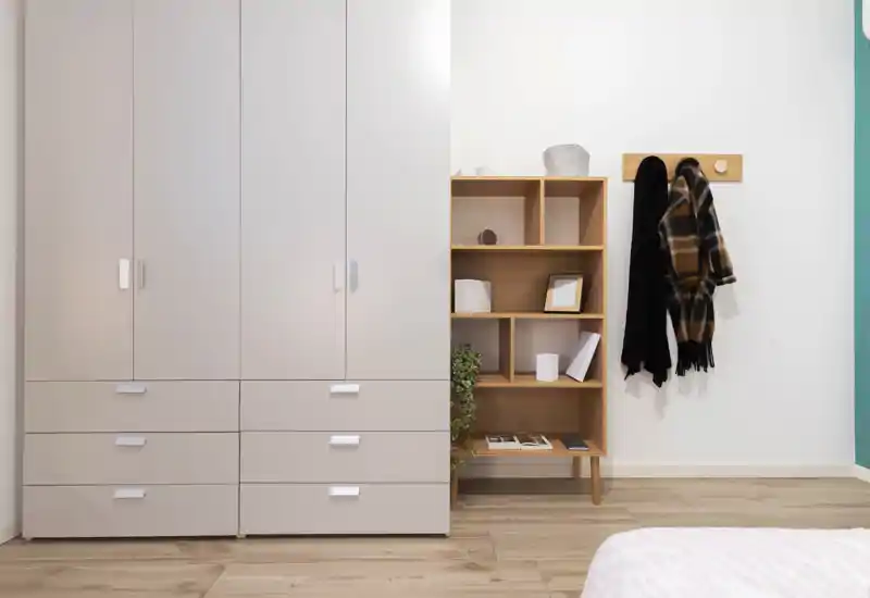 Bedroom corner with a large built-in wardrobe, an open wooden shelf and a partially visible bed in the foreground. Neutral tones, tidy décor and bright natural-looking light create a calm, contemporary sleeping area.