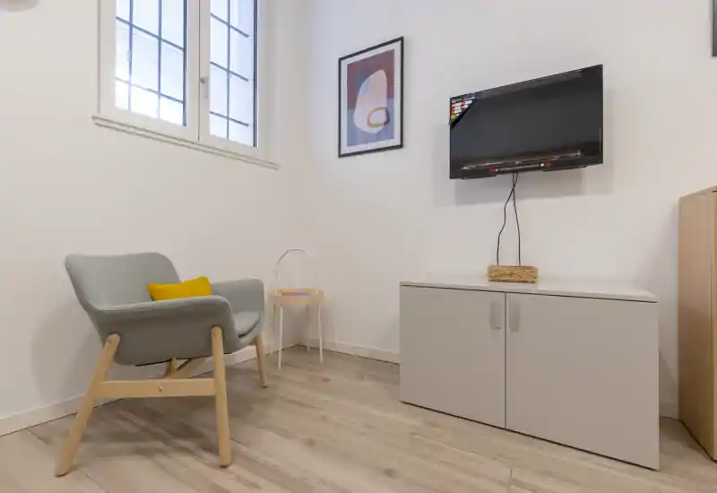 A small, modern living corner featuring a comfortable armchair with a accent cushion, a low cabinet with wall-mounted TV and bright natural light from a window — ideal for a cozy reading or relaxation nook.
