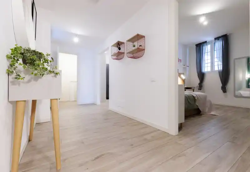 Modern apartment corridor leading to a bedroom: light wood floors, white walls, decorative shelving and a softly dressed bed visible through the doorway.