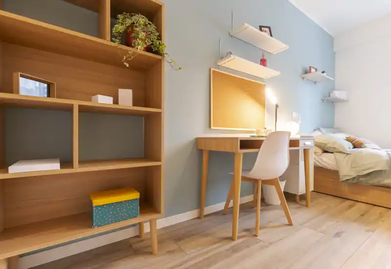 Bright, neatly staged bedroom with a single bed, wooden desk and shelving—ideal for a student or guest. The room features warm wood tones, soft bedding and a small workspace by the wall.