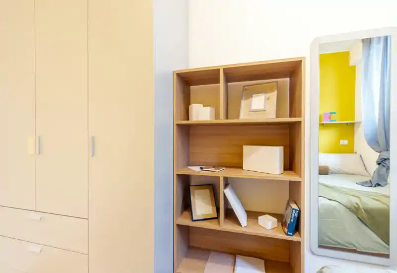 Compact bedroom corner with a light wood bookshelf, a tall wardrobe and a wall mirror reflecting part of the bed; bright, tidy and simply decorated.