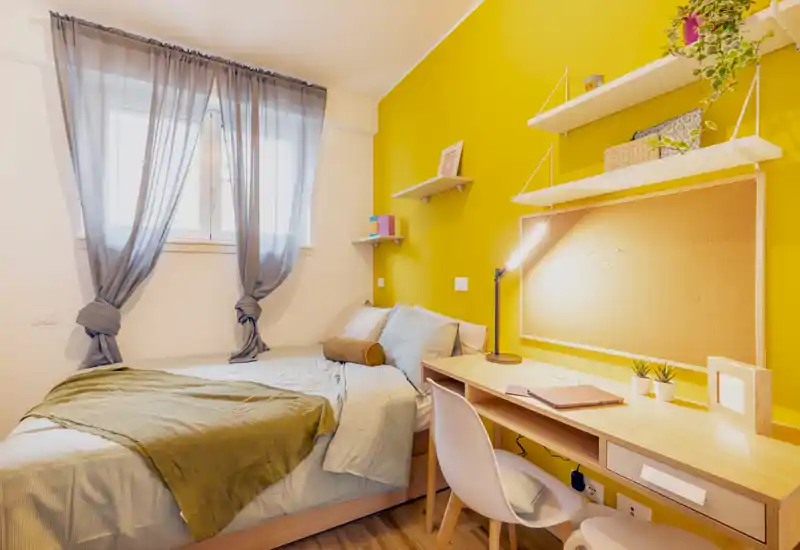 Sunny, compact bedroom with a full visible bed, cozy linens and a functional study desk by the wall — bright yellow accent wall adds personality.