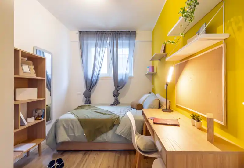 Bright, well-styled single bedroom with a comfortable bed, desk workspace and warm yellow accent wall — ideal for a student or young professional.