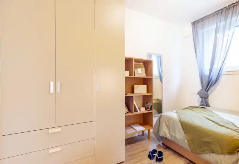 Bright, tidy bedroom with a double bed, soft green throw and natural light from a curtained window; built-in wardrobe and shelving provide storage.