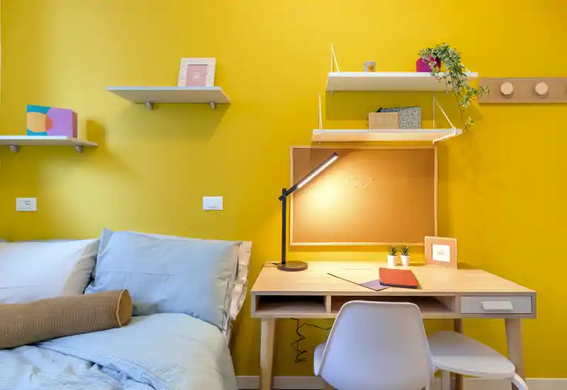 Bright, well-styled bedroom corner with a comfortable bed and a tidy study desk against a warm yellow wall. Ideal image for listings highlighting a cozy sleeping area with a workspace.