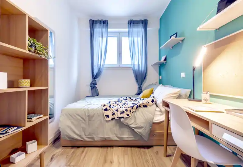 Bright, well-styled single bedroom with a comfortable bed placed under a window, a wooden desk with task lighting and shelving — ideal for a student or solo professional.
