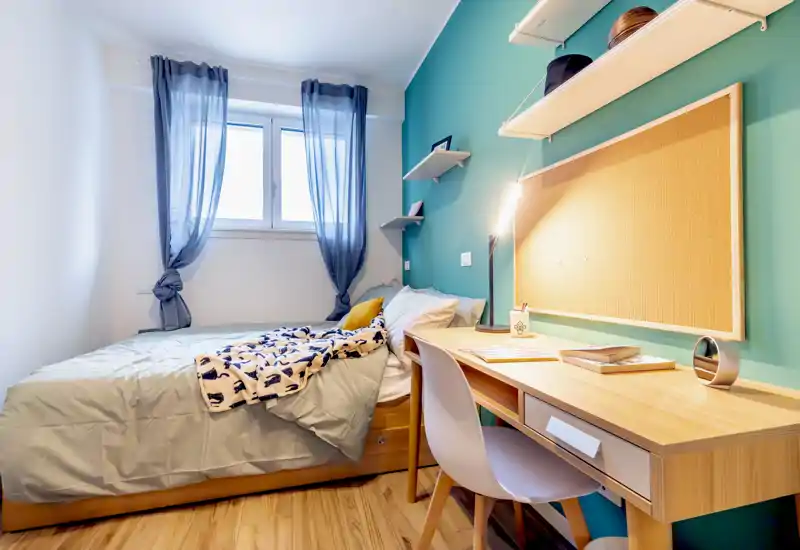 Compact, well-lit single bedroom with a comfortable bed, bedside shelves and a wooden study desk with lamp — ideal for students or a single occupant.