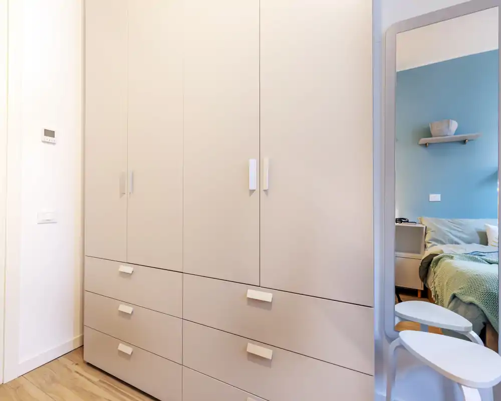 Vista di una camera moderna con focus sull'ampio armadio a muro; il letto è visibile solo parzialmente nello specchio sul lato destro. La scena è luminosa e nitida, con toni neutri e pavimento in legno.