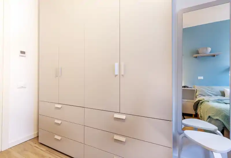 Vista di una camera moderna con focus sull'ampio armadio a muro; il letto è visibile solo parzialmente nello specchio sul lato destro. La scena è luminosa e nitida, con toni neutri e pavimento in legno.