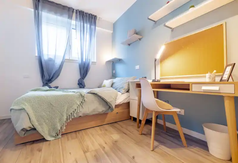 Camera singola luminosa e accogliente con letto ordinato, mensole sopra il comodino e una scrivania in legno compatta, perfetta per studiare o lavorare da casa.