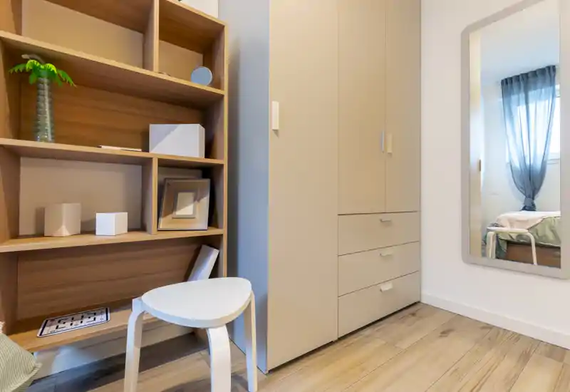 Small, well-lit bedroom corner shown with wardrobe, shelving and a mirror reflecting the bed and window; clean modern styling and light wood floors create a fresh, airy feel.