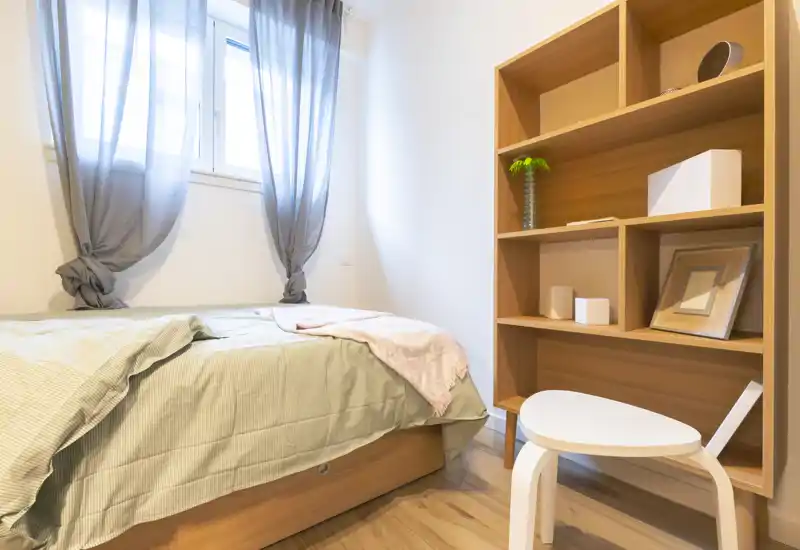 Bright, simply styled bedroom with a double bed, soft linen and a wooden shelving unit; natural light from the window creates a calm, airy atmosphere ideal for rental photos.