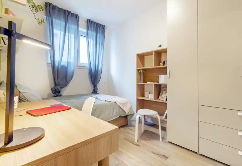 Bright, cozy single bedroom with a study desk, shelving and wardrobe. Soft natural light through sheer curtains and warm wood tones create a comfortable atmosphere ideal for students or professionals.