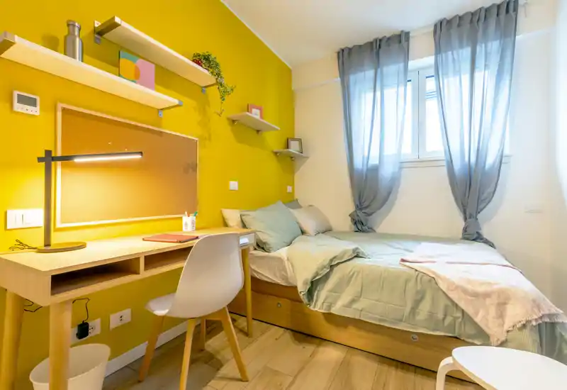 Bright, cozy single bedroom with a wooden bed, soft pastel bedding and a study desk by a warm yellow accent wall. The window provides natural light while sheer curtains add a light, airy feel — ideal for a comfortable studio or student room.