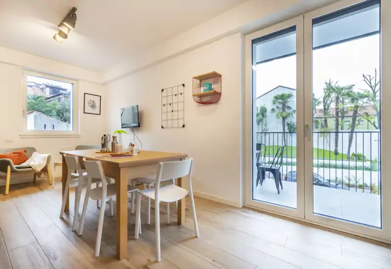 Bright, modern dining area with a wooden table and four chairs next to large glass doors opening to a small balcony. Natural light and warm wood flooring create an inviting atmosphere perfect for meals or working.