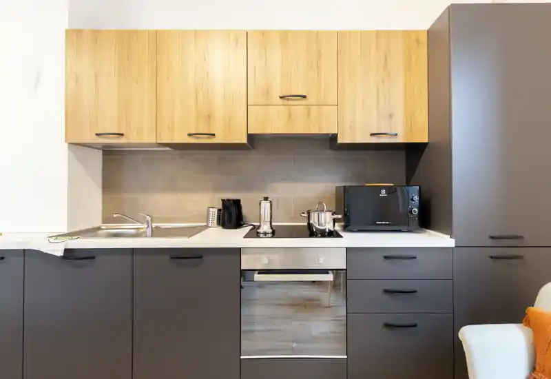 Modern compact kitchen with wooden upper cabinets, dark lower units, integrated oven and countertop appliances. Bright, clean layout ideal for rental listings highlighting functionality.