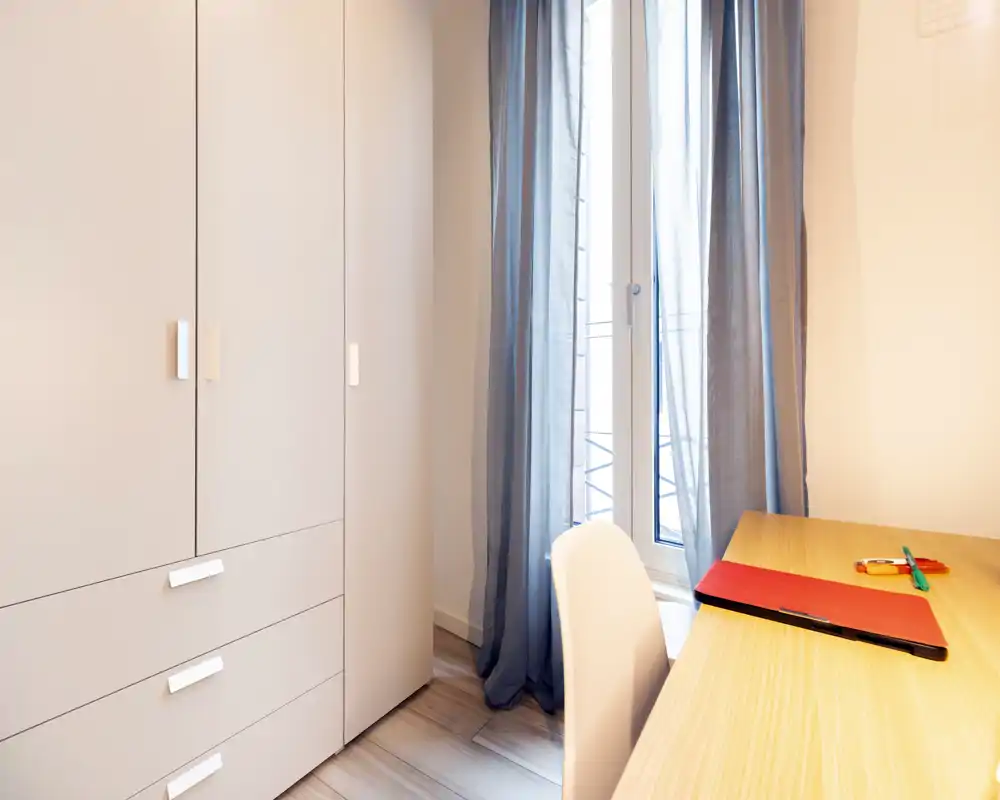 Coin chambre lumineux et minimaliste avec une grande armoire, un bureau en bois et une fenêtre avec voilages — propre, moderne et bien éclairé.