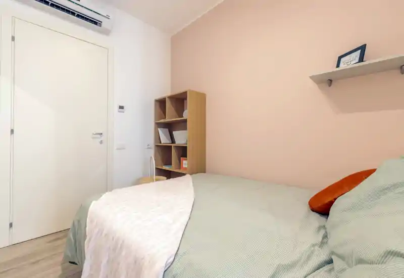 Dormitorio luminoso y acogedor con cama doble bien hecha, tejidos suaves y paredes en tonos pastel; estantería compacta y decoración cuidada para una estancia cómoda.