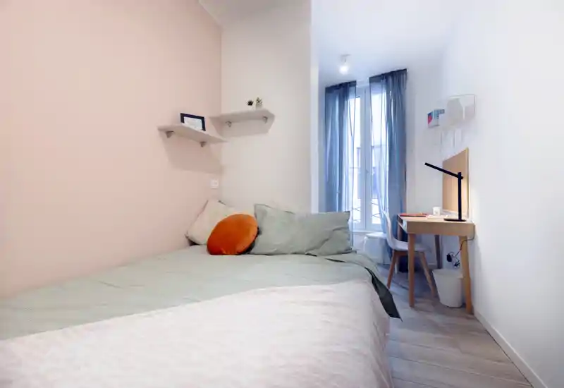 Dormitorio luminoso y compacto con cama bien hecha, cojines decorativos y una pequeña zona de trabajo junto a la ventana — ideal para una persona o estancias cortas.