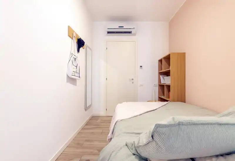 Dormitorio compacto con cama individual junto a la pared, pared de acento en tonos pastel, estantería abierta y aire acondicionado sobre la puerta. Luminoso, minimalista y bien preparado para un anuncio de alquiler.