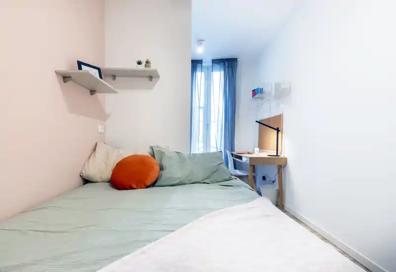 Dormitorio compacto y luminoso con cama bien hecha, cojines cómodos y un pequeño escritorio junto a la ventana. Estilo minimalista y luz natural, ideal para alquileres de corta estancia o estudiantes.