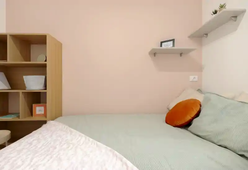 Dormitorio luminoso de estilo suave con cama bien hecha, cojines decorativos y estanterías minimalistas: ideal como habitación individual o de invitados.