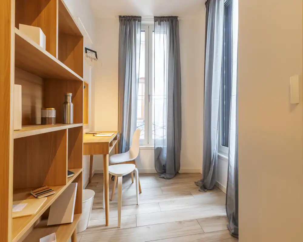Stanza luminosa e minimal con una libreria in legno e una scrivania compatta davanti a finestre alte — ideale come angolo studio o piccolo soggiorno.
