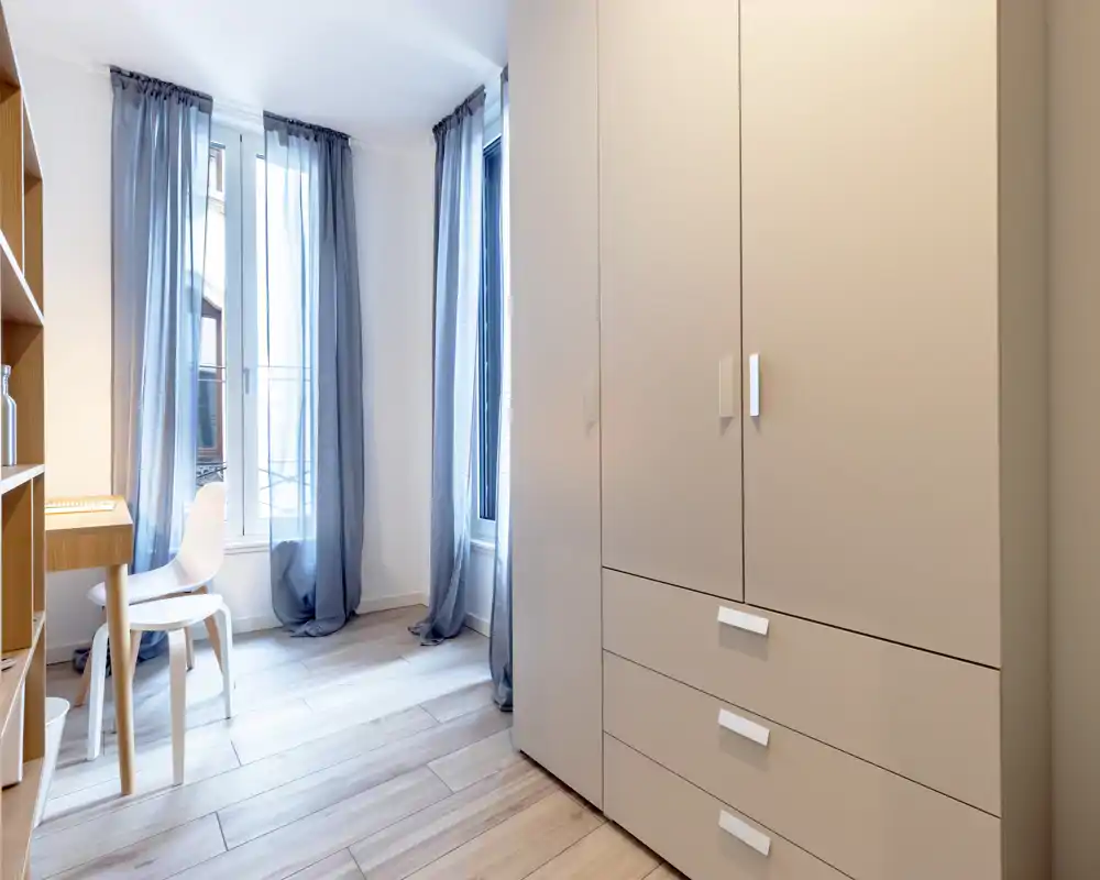 Angolo di camera da letto moderno e luminoso con grande armadio, luce naturale da ampie finestre e una piccola scrivania vicino alla finestra — ideale per una camera compatta o monolocale.