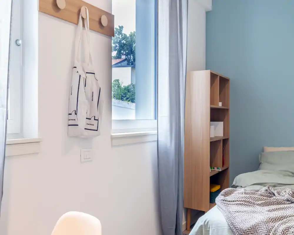 Bright, simply styled bedroom corner with a neatly made bed, wooden shelving and natural light from a window; calm pastel tones and soft textiles create a relaxed atmosphere.