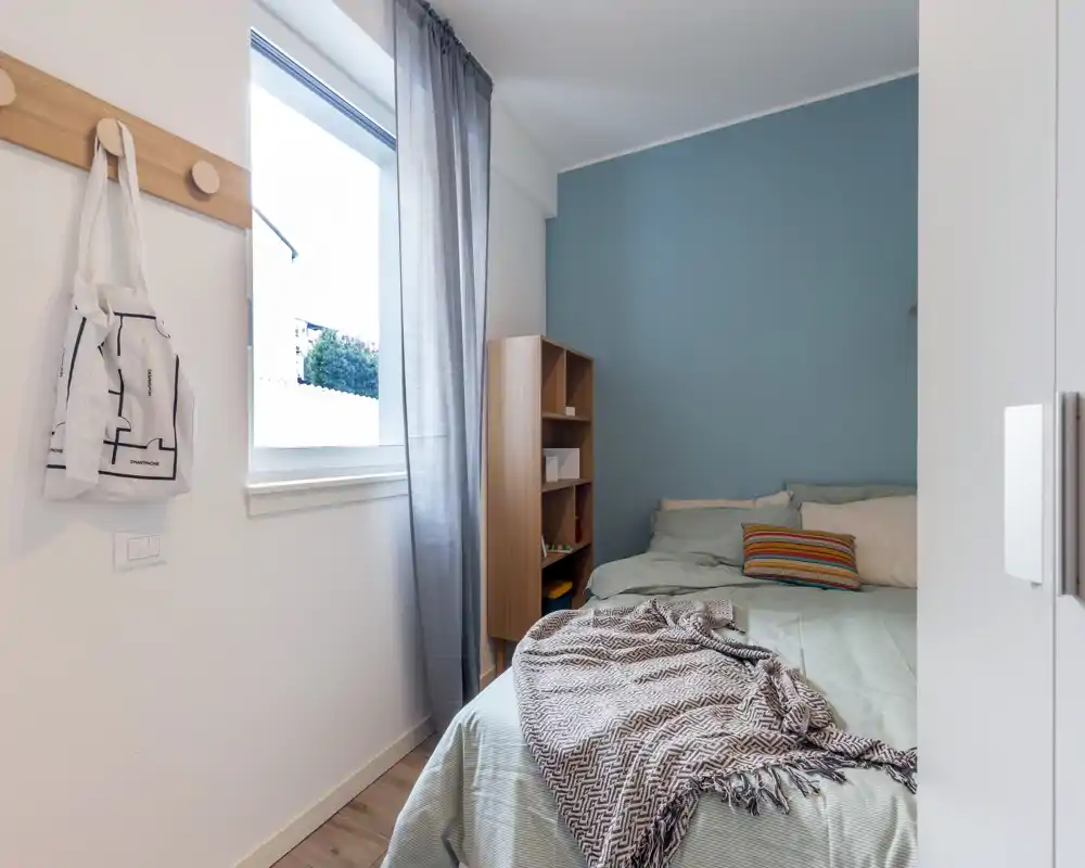 Compact, well-lit bedroom with a double bed dressed in soft linens, a textured throw and a colorful cushion. Natural light pours through the window and a slim shelving unit provides practical storage.