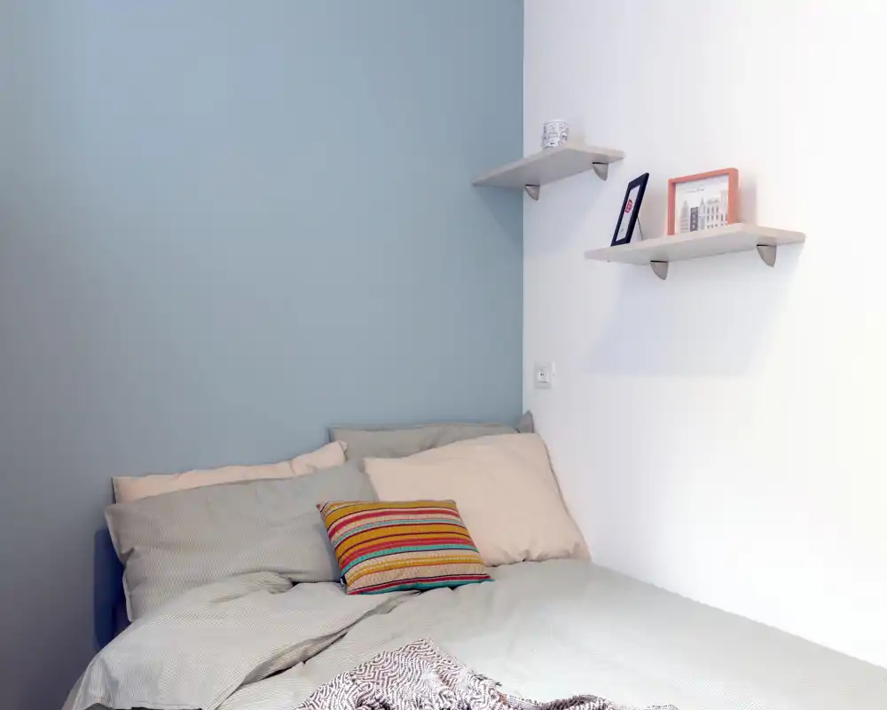 Bright and calm bedroom corner with a neatly made double bed, soft pillows and a colorful accent cushion against a pale blue wall.