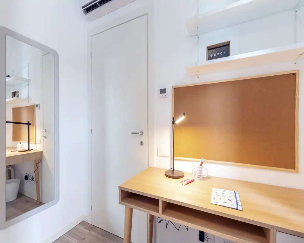Small, bright bedroom corner with a wooden desk, corkboard and a full-length mirror reflecting a tidy workspace; simple, modern and well lit.