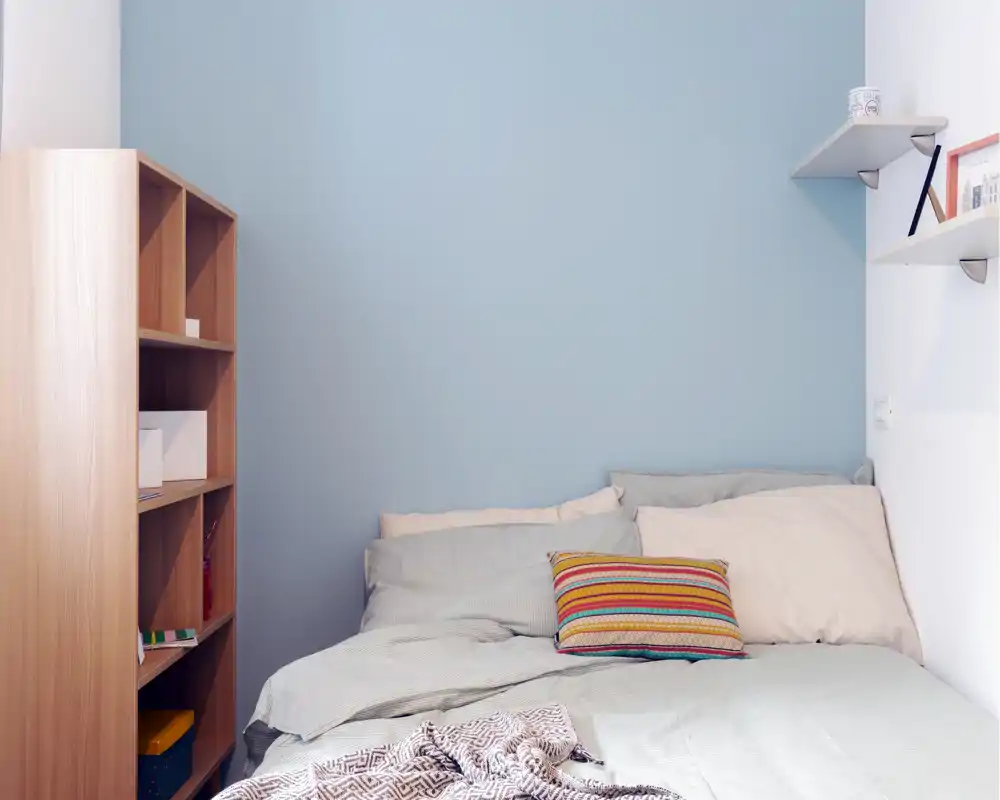 Small, cozy bedroom with a neatly made bed, layered linens and a colorful decorative cushion against a pale blue wall; light wooden shelving provides storage and styling.