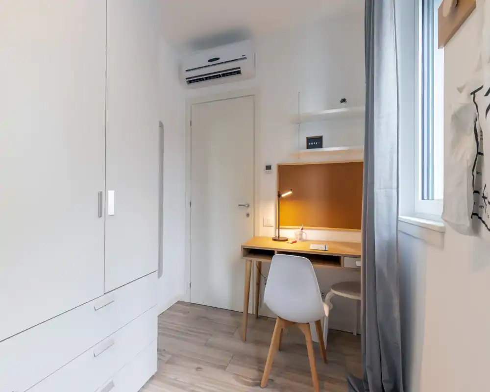 Compact bedroom corner with a study desk, wardrobe and window; bright natural light and a warm desk lamp create a clean, functional atmosphere.