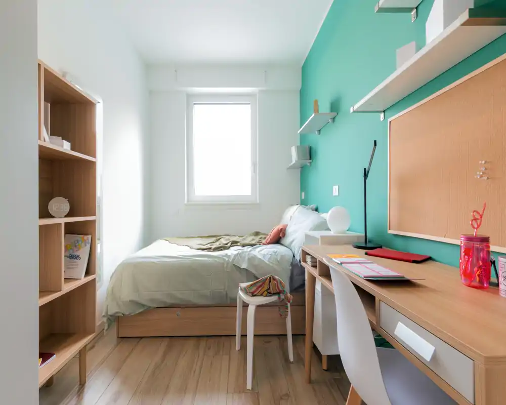 Bright, well-styled single bedroom with a comfortable bed, wooden floor and a study desk alongside an accent teal wall. Natural light from the central window makes the room airy and suitable for a rental listing.