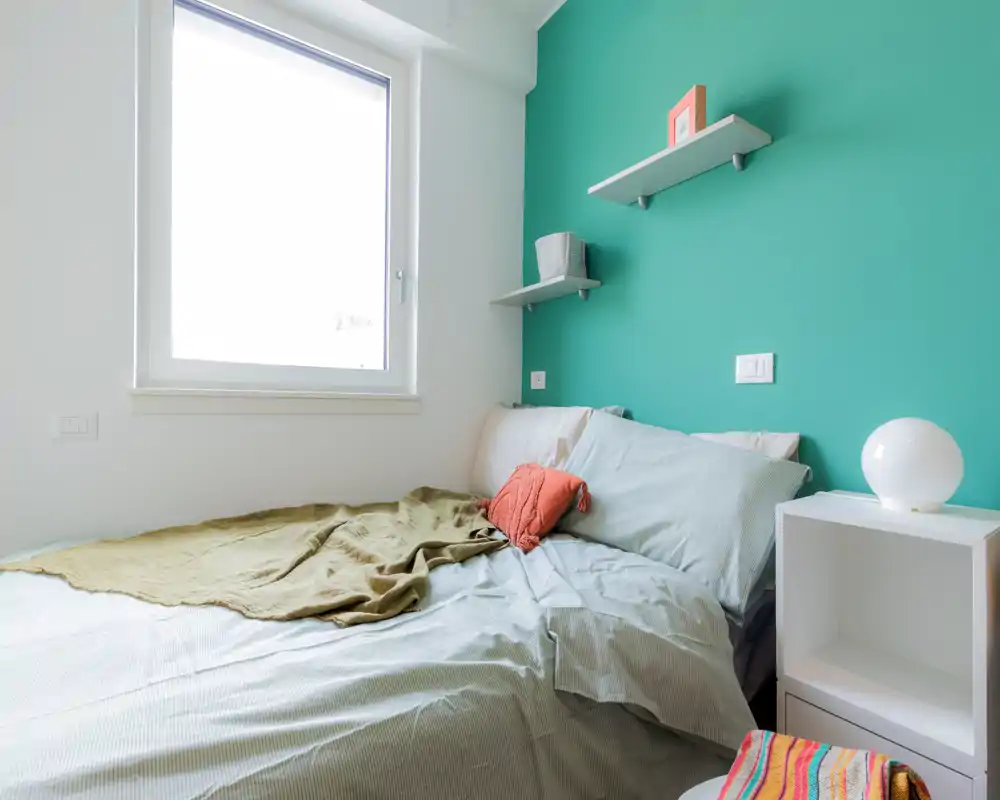 Bright, cozy bedroom with a teal accent wall, neatly made double bed, bedside shelf and natural light from a large window.