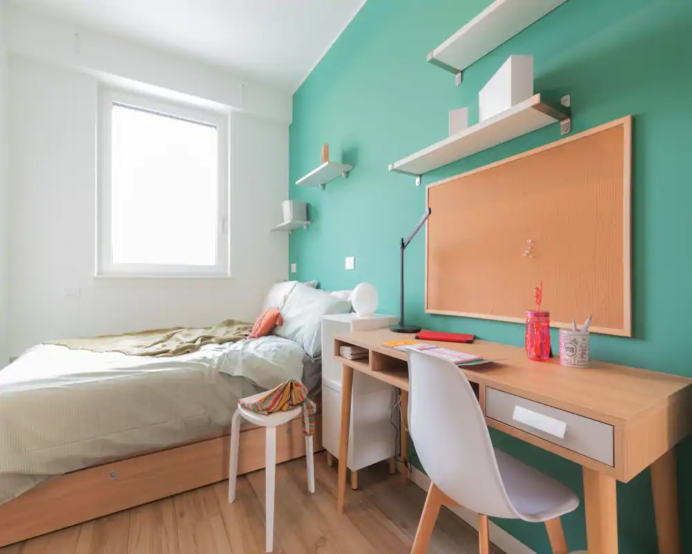 Bright, well-staged single bedroom with a compact study desk, wall shelves and a comfortable bed beneath a large window. Clean Scandinavian-style décor with green accent wall and wooden flooring.