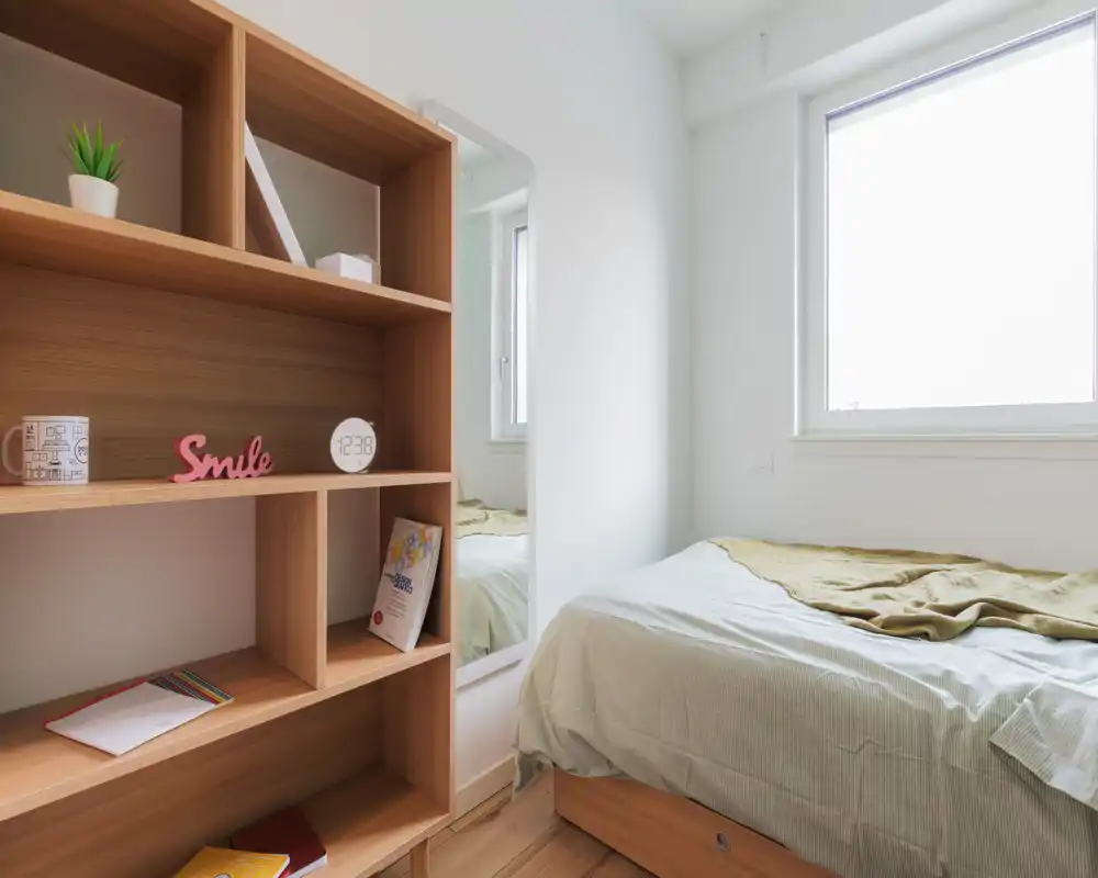 Bright, minimal bedroom with a neatly made single bed beneath a large window and a wooden shelving unit with decorative items.