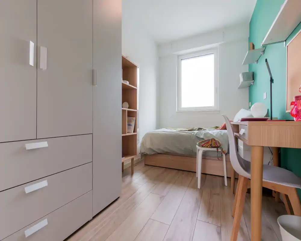 Neat, well-lit bedroom with a single bed positioned under a window, wooden floor and practical desk — ideal for a student or single occupant.