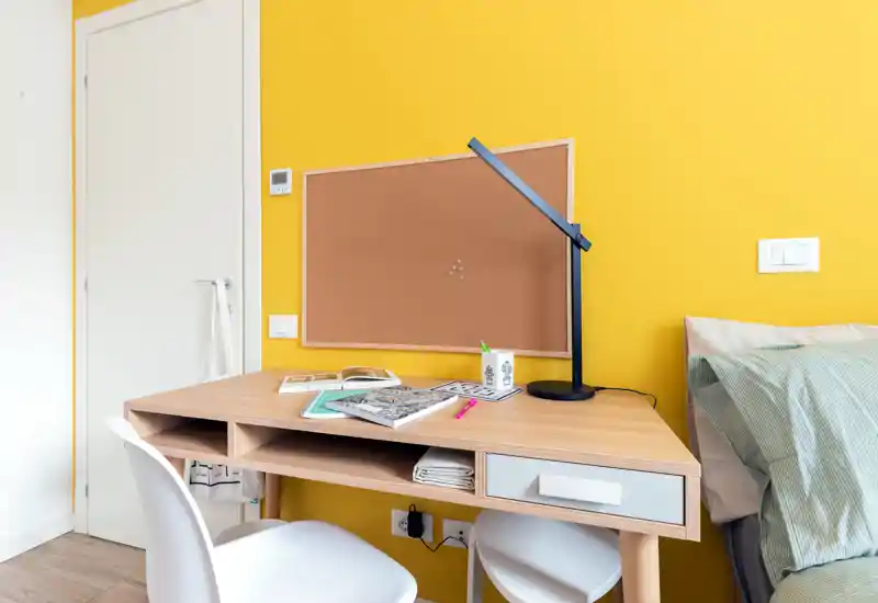 Bright, modern bedroom corner with a wooden study desk, cork notice board and part of the bed visible to the right—ideal for a student or home-office setup.