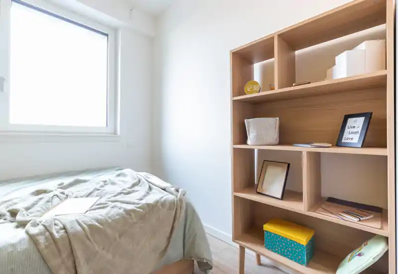Bright, minimal bedroom with a neatly made single bed beside a large window and a wooden shelving unit with decorative items.