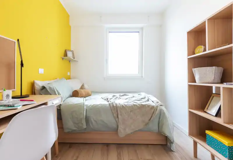 Bright, modern single bedroom with a neatly made bed, light wooden furniture and a yellow accent wall that adds warmth.