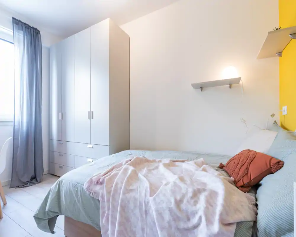 Bright, simply styled bedroom with a comfortable double bed, soft linens and a large white wardrobe by the window. The light and textiles create a welcoming, modern feel ideal for property listings.